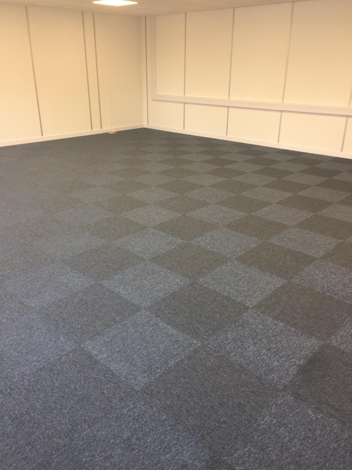 A finished commercial carpet-tile floor laid wall to wall, the tiles alternating light and dark in a chequered pattern, against panelled walls